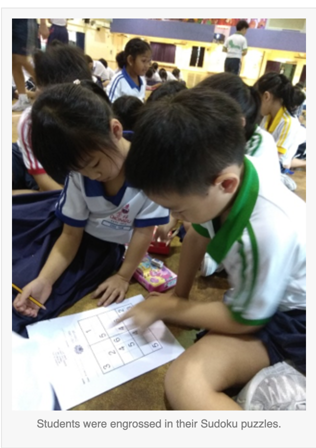 Two young students lean over a Sudoku puzzle worksheet on the floor in a busy school hall.